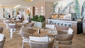 Een modern restaurant met rieten meubels, een centraal gelegen bar en een muurschildering van tropische bladeren.