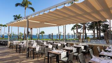 An outdoor dining area with tables and chairs, palm trees, and a view of the ocean.