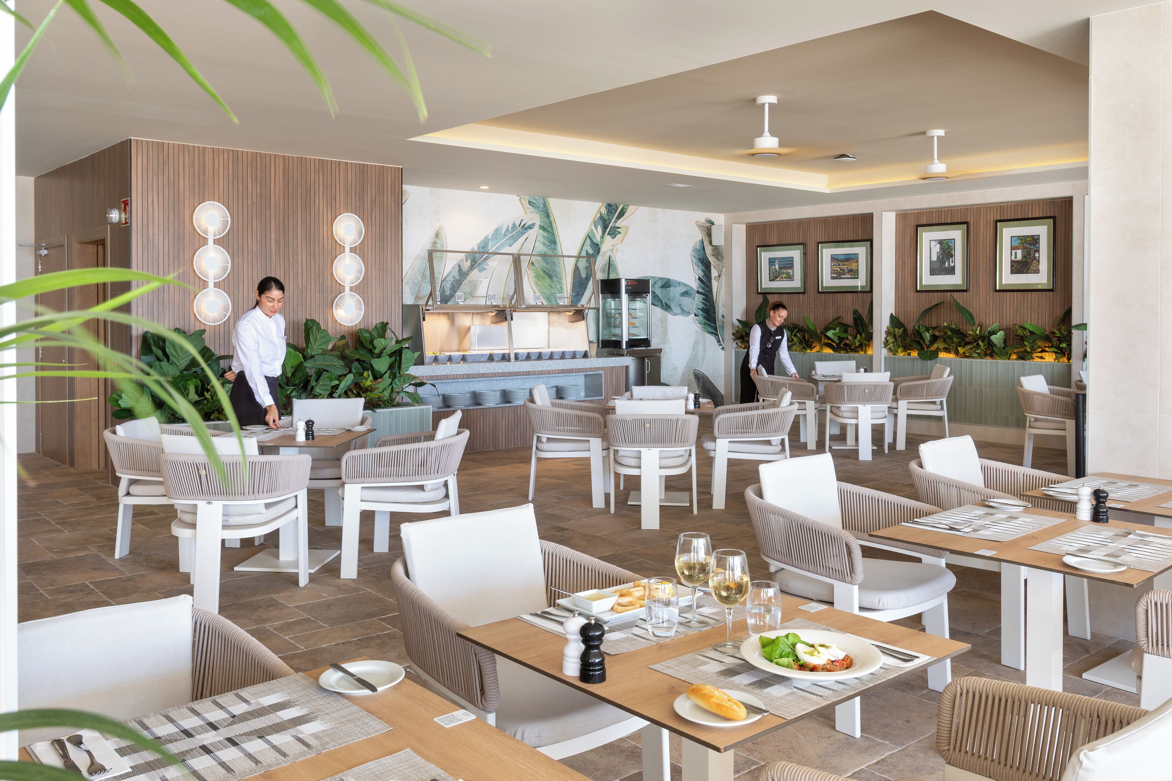 Ein modernes Restaurant mit einer zentralen Bar, verschiedenen Sitzbereichen und einem tropischen Wandbild.