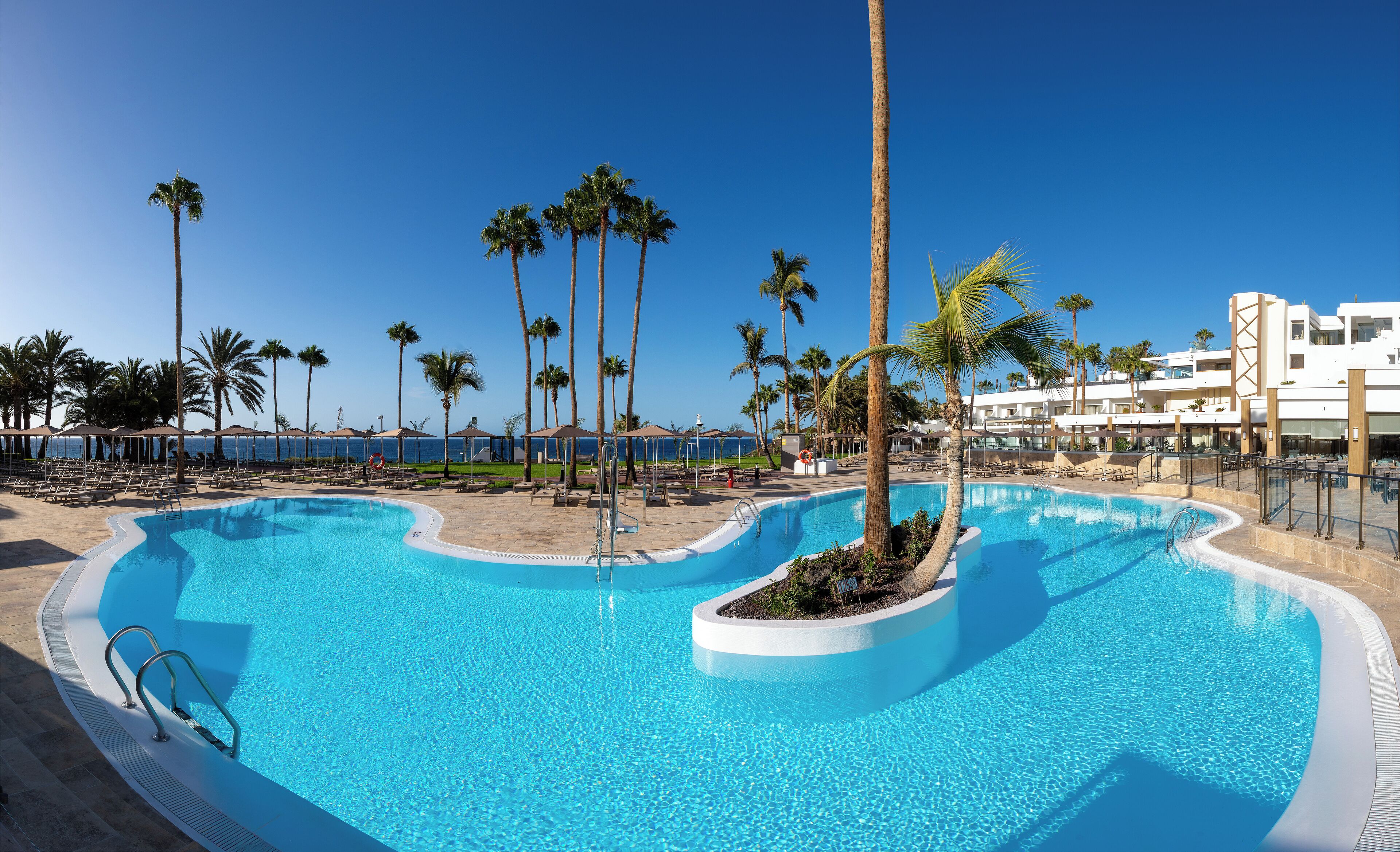 Zona de piscina con agua cristalina y color turquesa, rodeada de palmeras y sillas de descanso, con vista al océano y un edificio al fondo.