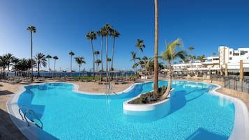 Zona de piscina con agua cristalina y color turquesa, rodeada de palmeras y sillas de descanso, con vista al océano y un edificio al fondo.