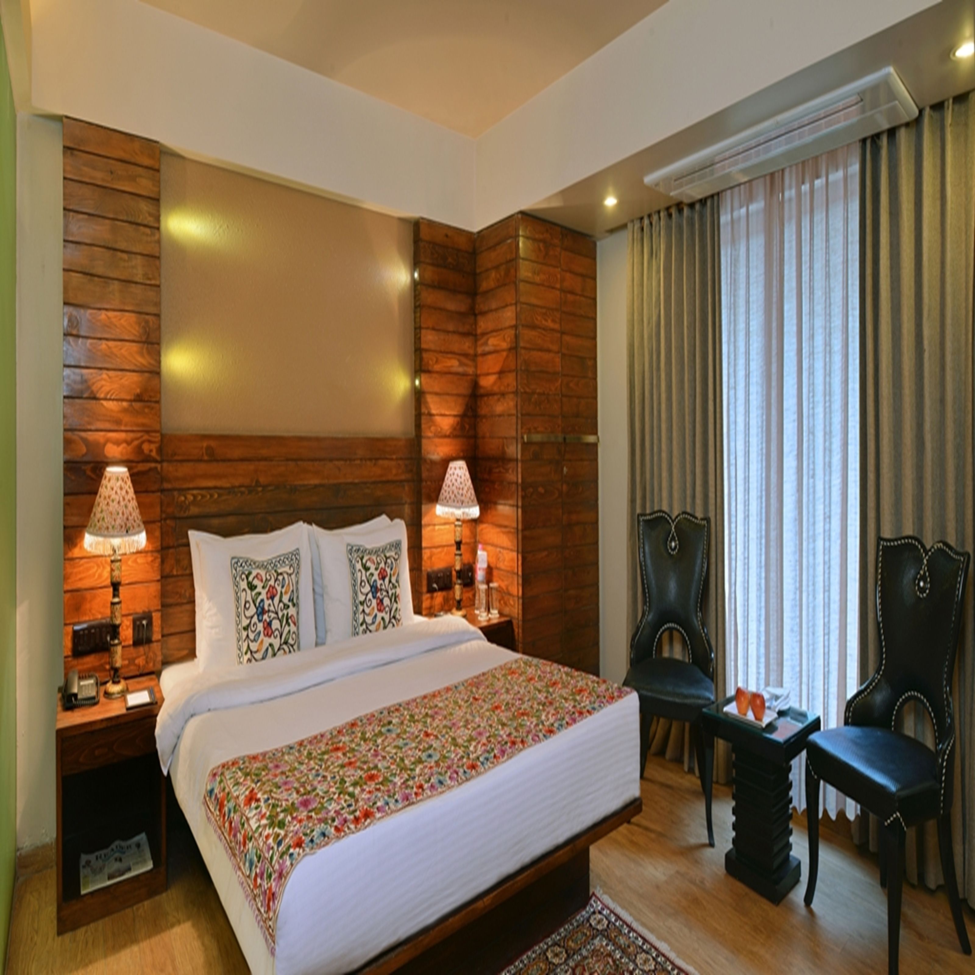 A hotel room with a wooden headboard, a large bed with floral bedding, two bedside lamps, a chair, a small table, and a window with curtains.