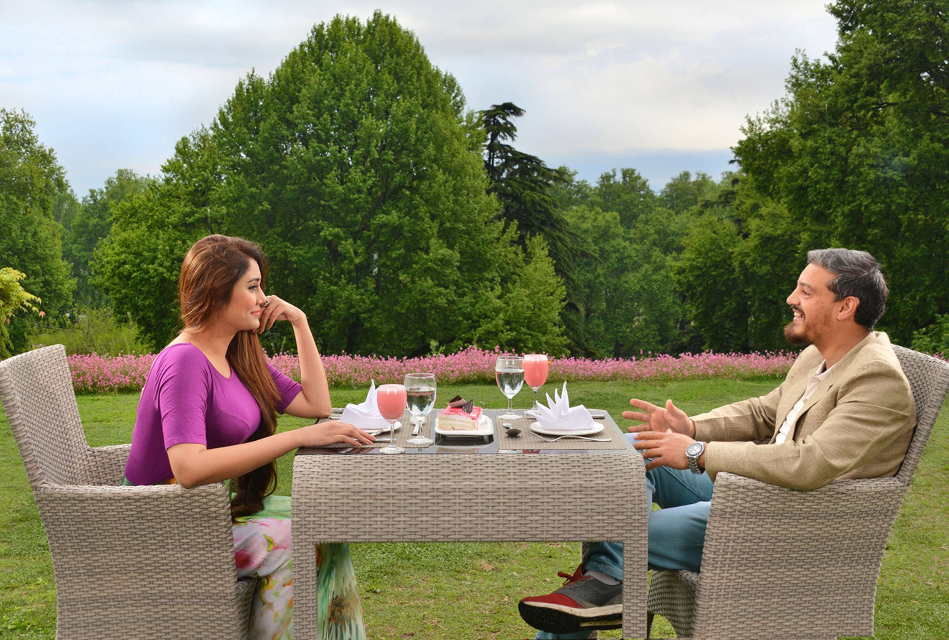 Two people seated at an outdoor table with a cake and drinks, surrounded by greenery.