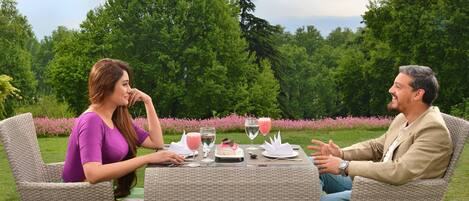 Two people seated at an outdoor table with a cake and drinks, surrounded by greenery.