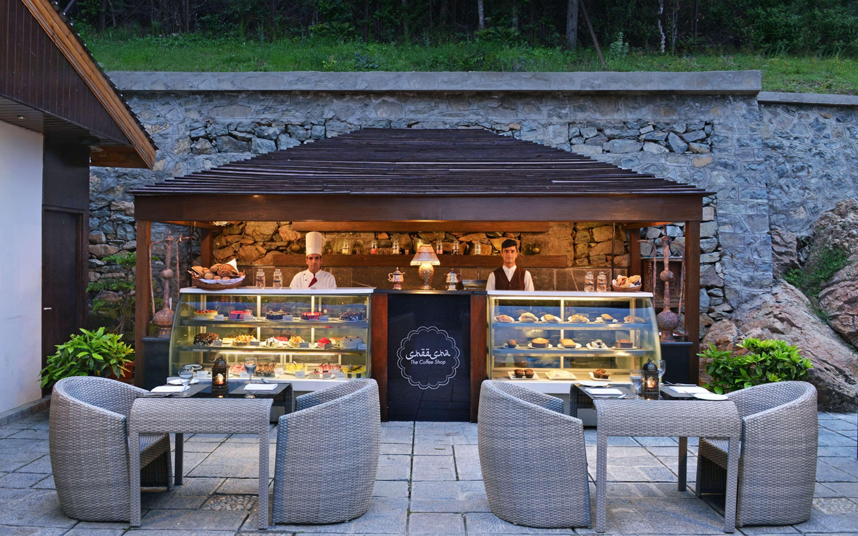 Un café al aire libre con mobiliario de mimbre, un muro de piedra de fondo y una vitrina con pasteles.