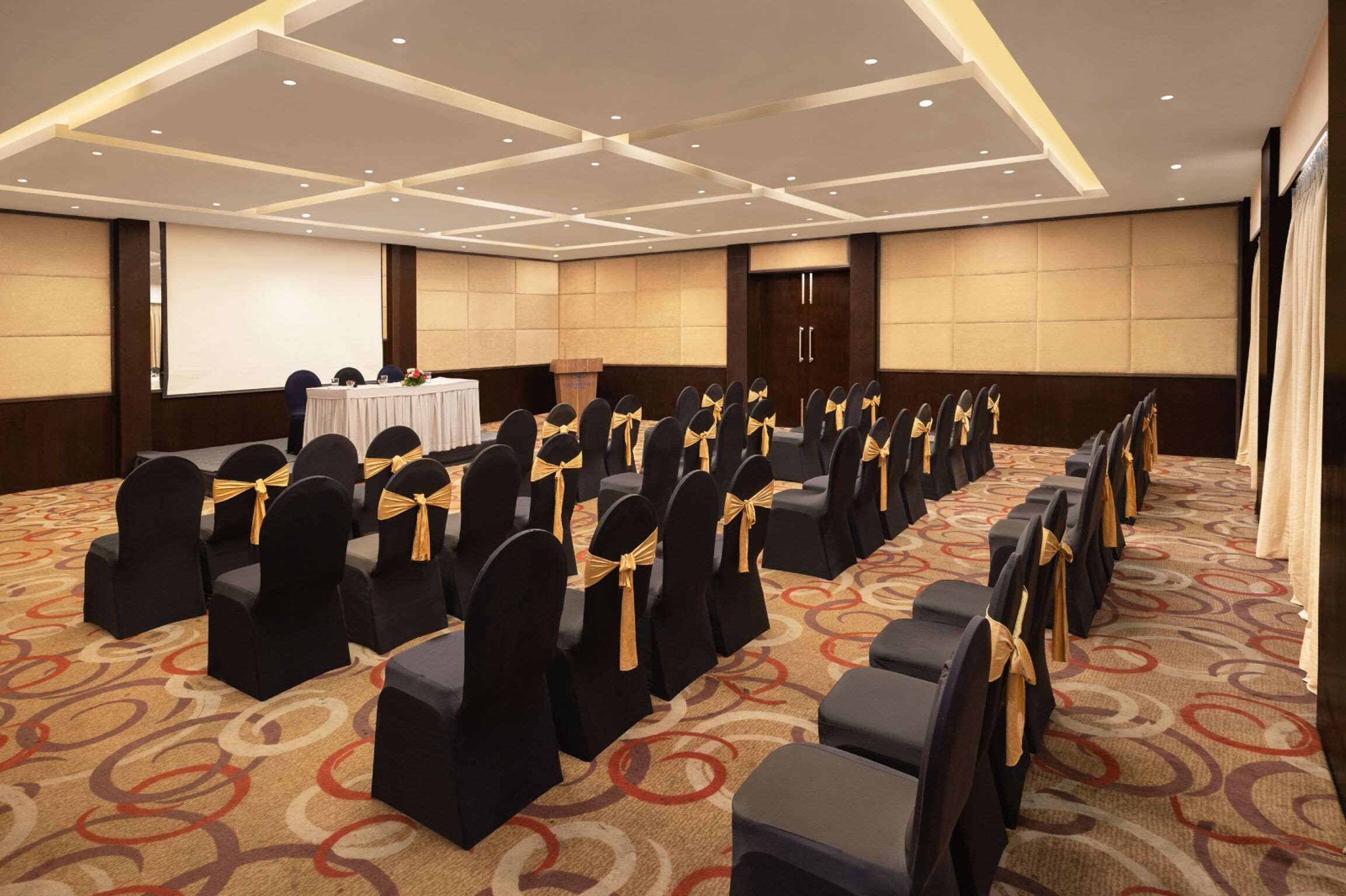 A conference room with rows of black chairs with gold sashes, a podium, and a projector screen.