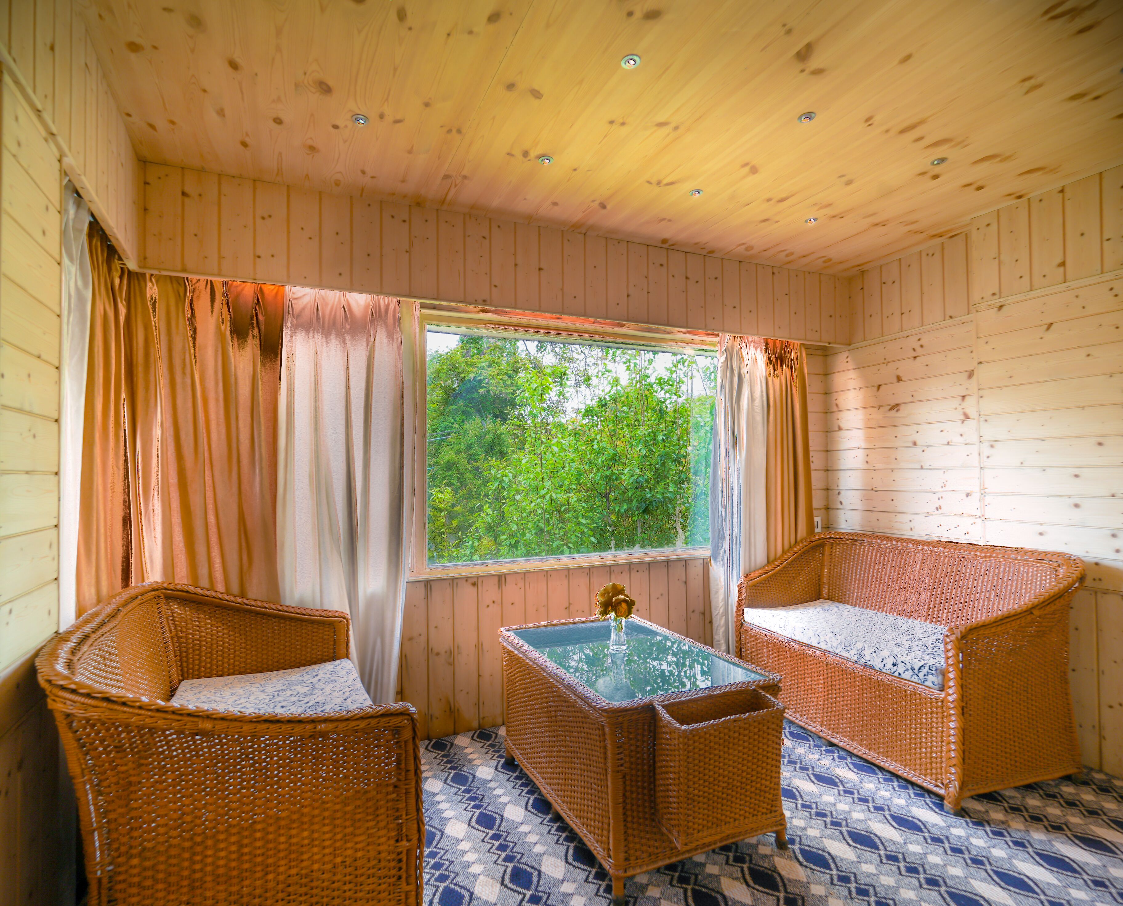 A room with wooden walls and ceiling, two wicker chairs, a glass-top table, and a window with curtains.