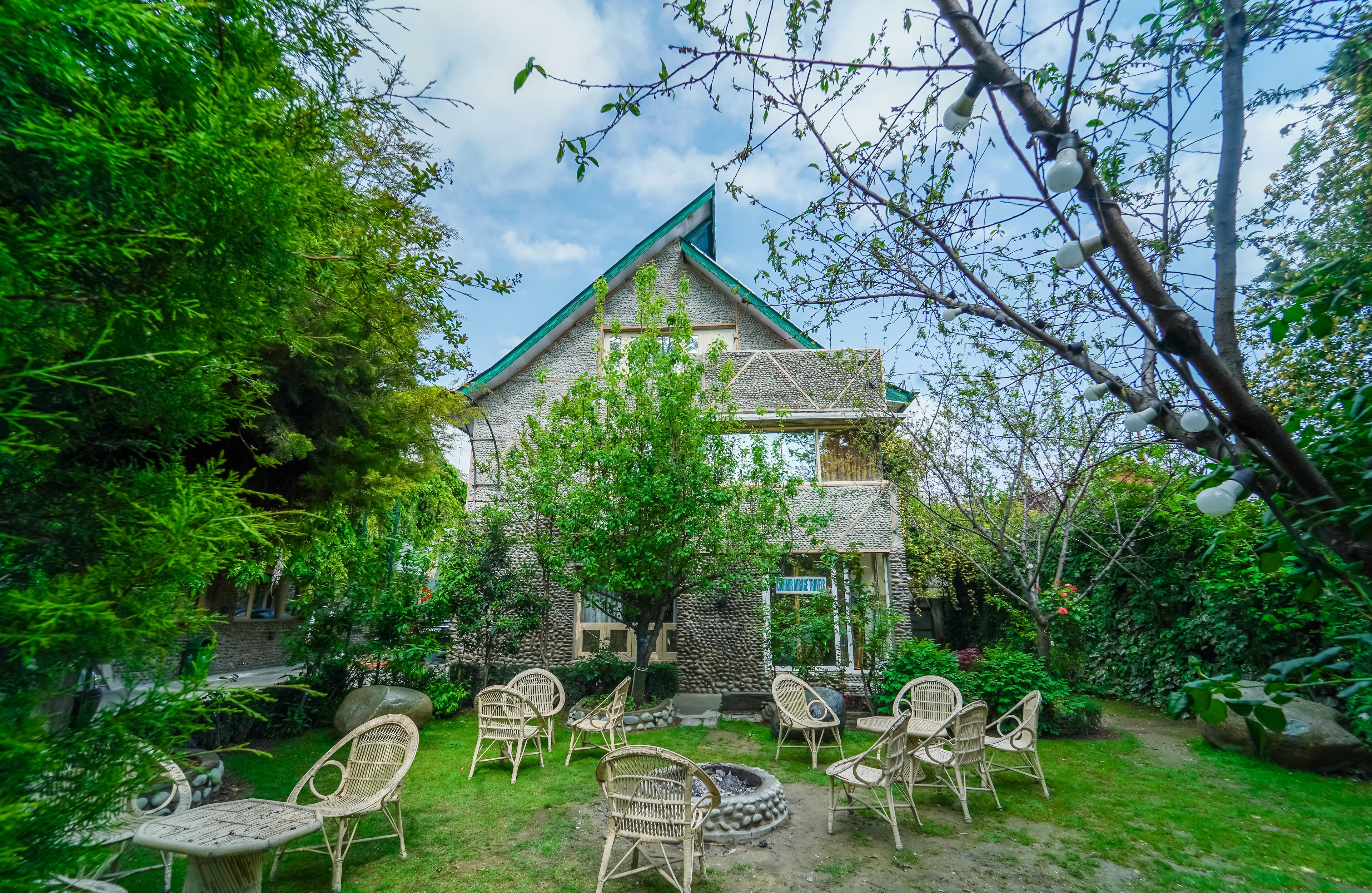 Un edificio de piedra con techo verde, rodeado de abundante vegetación y mobiliario exterior.