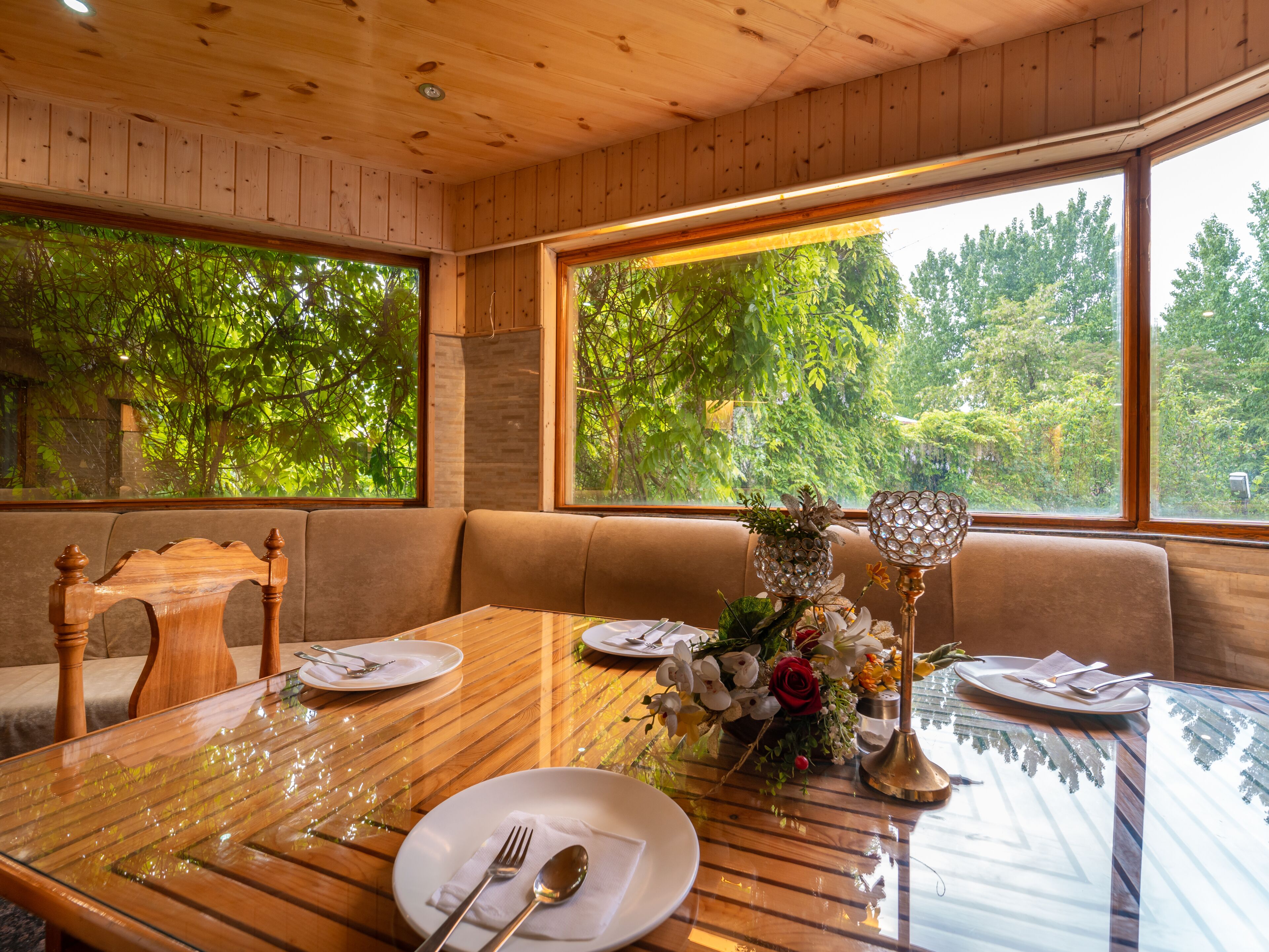 A dining area with a wooden table, glass top, and a view of trees through large windows.