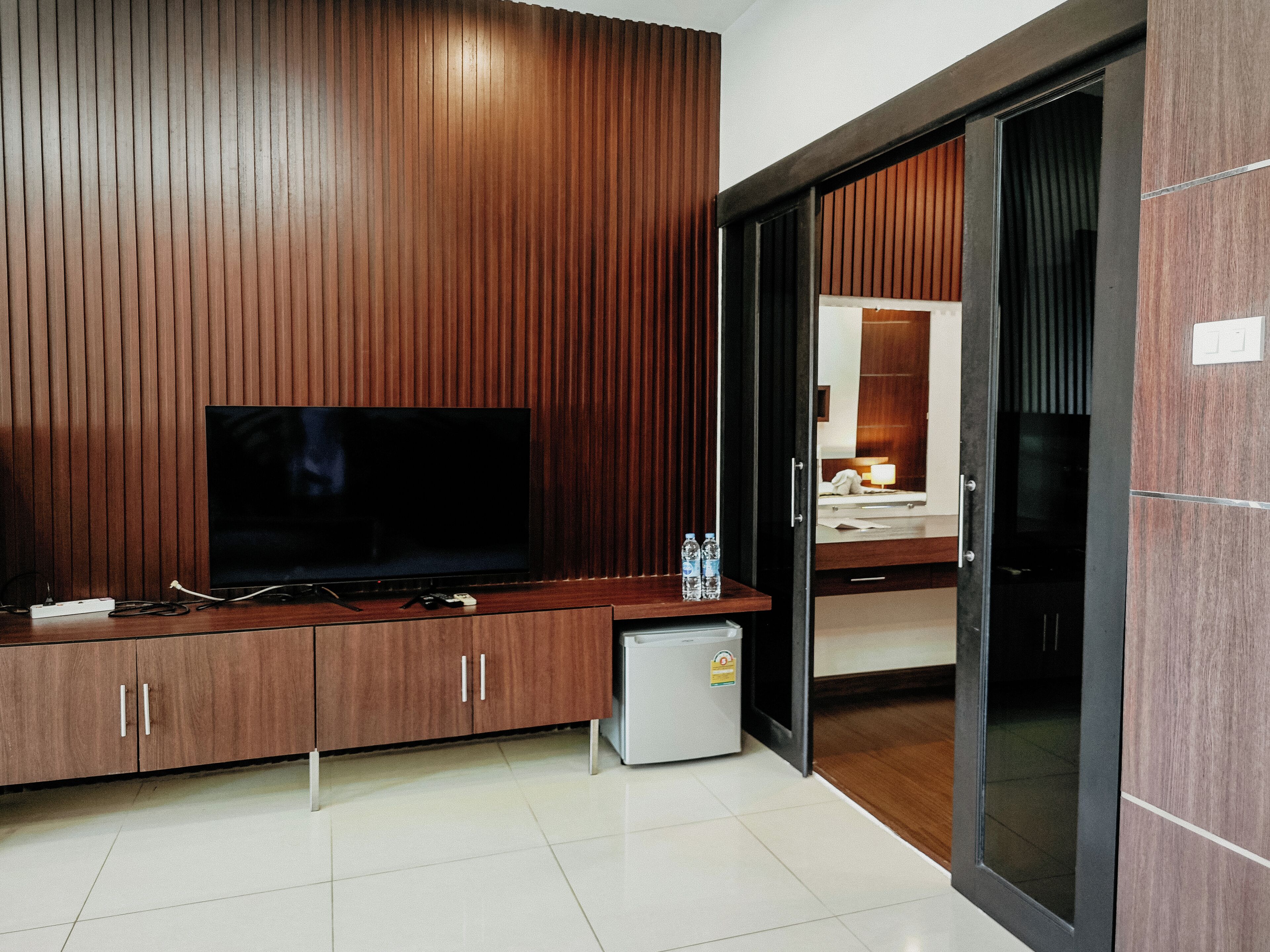A modern hotel room with a wooden panel wall, a flat-screen TV, a mini-fridge, and a small table with two bottles of water.
