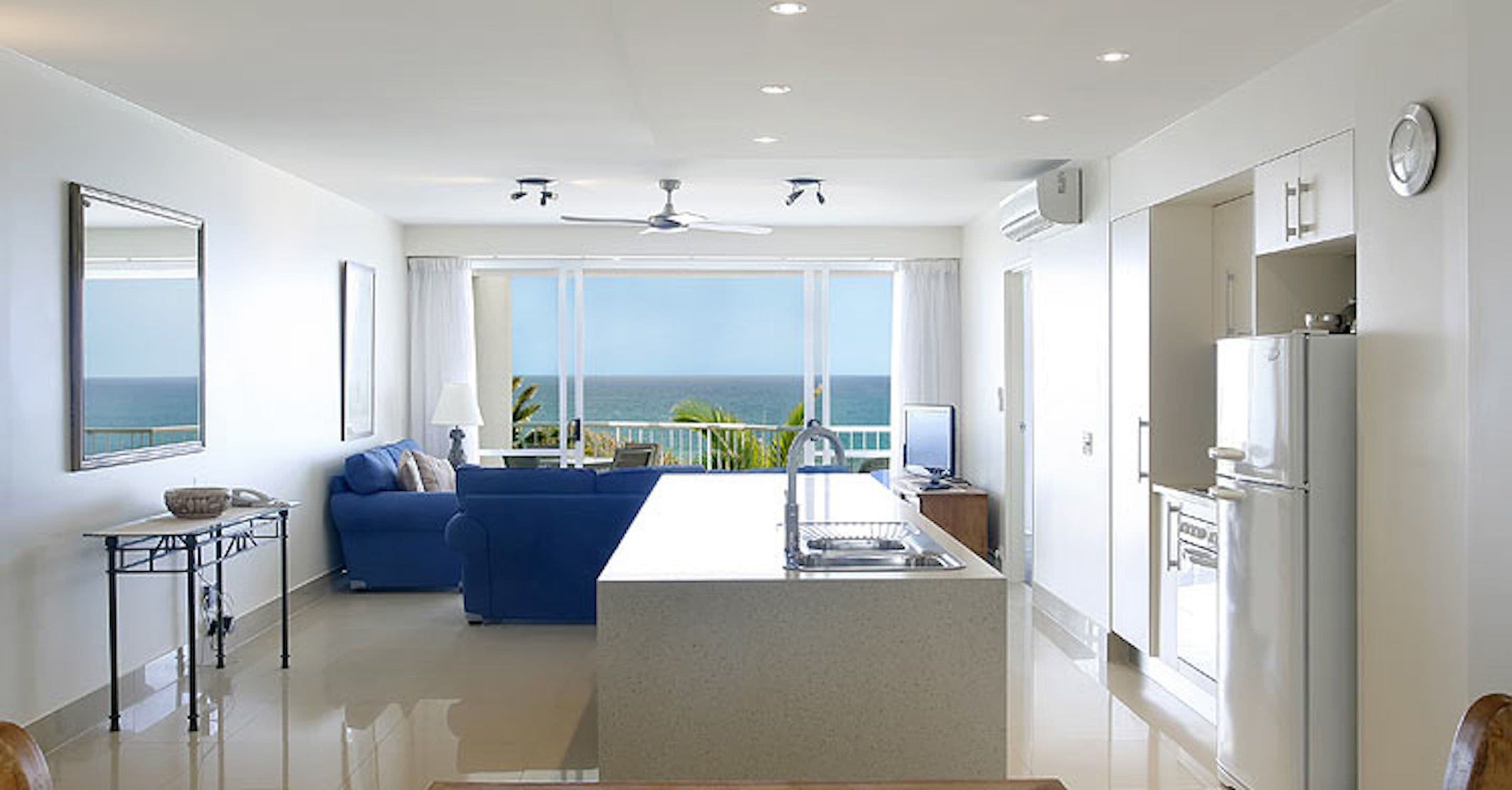 A modern kitchen with a central island, a dining area with a blue sofa, and a view of the sea.