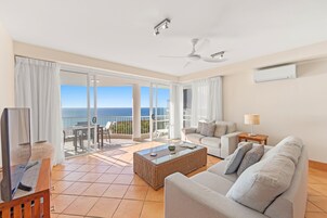A spacious living area with a large sliding glass door leading to a balcony with a view of the sea. There is a comfortable sofa, a coffee table, and a television.