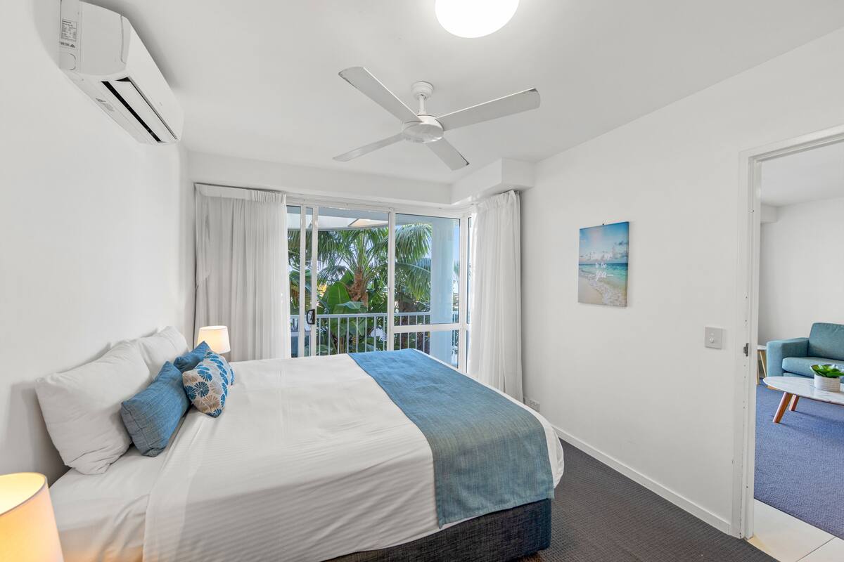 Ein Schlafzimmer mit einem Bett, Deckenventilator, Klimaanlage, einem Fenster mit Vorhängen und Blick ins Freie.