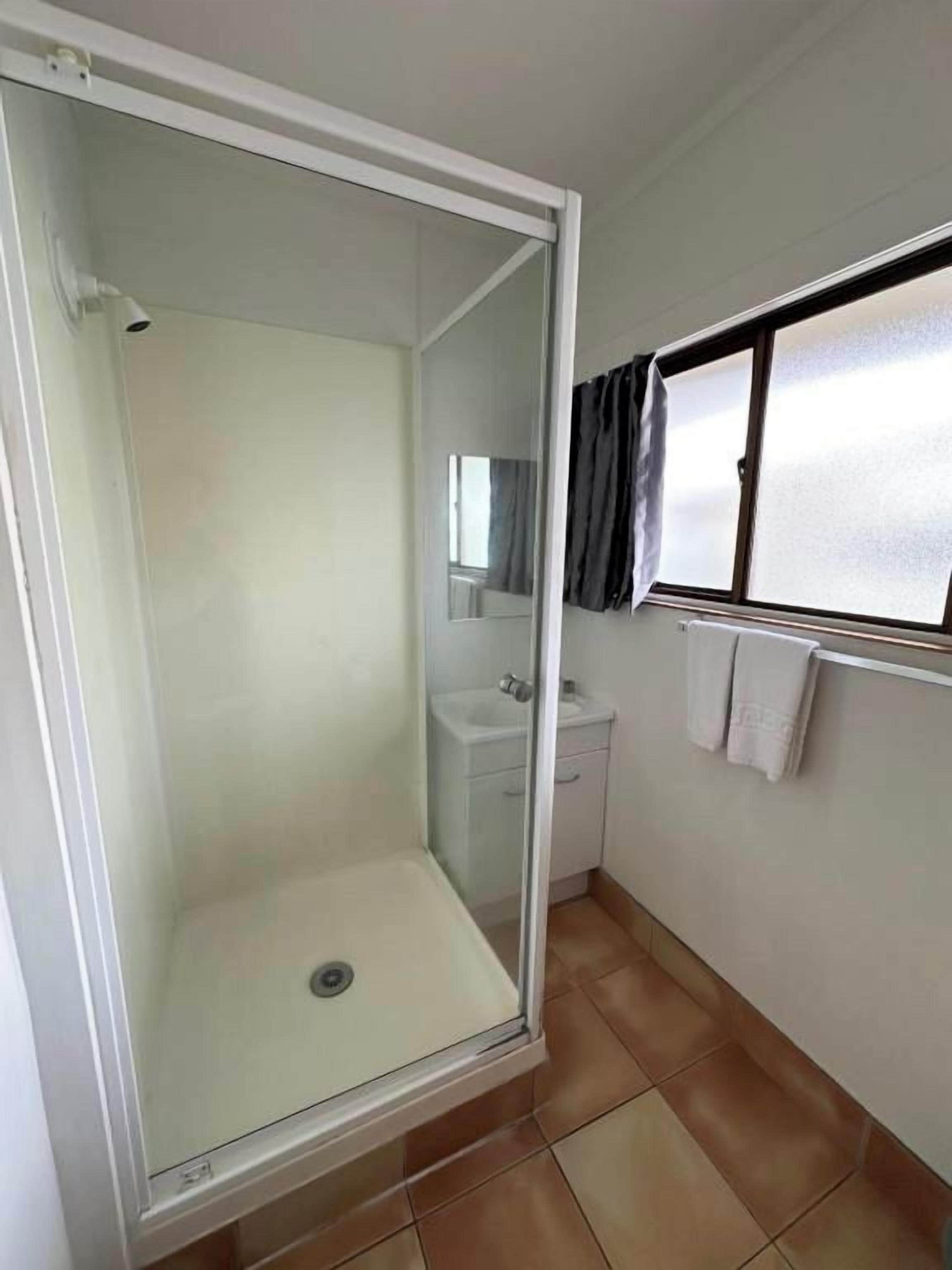 Bany amb dutxa trencada per vidre, un lavabo i una finestra amb cortines.