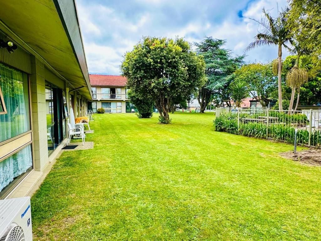Gramado bem cuidado com uma grande árvore ao centro, um edifício com varanda à esquerda e uma área cercada com plantas à direita.