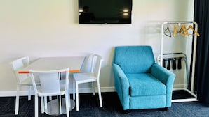 A hotel room with a flat-screen TV mounted on the wall, a blue armchair, a small table with two white chairs, and a coat rack with hangers. - Airport Gold Star Motel (Auckland)