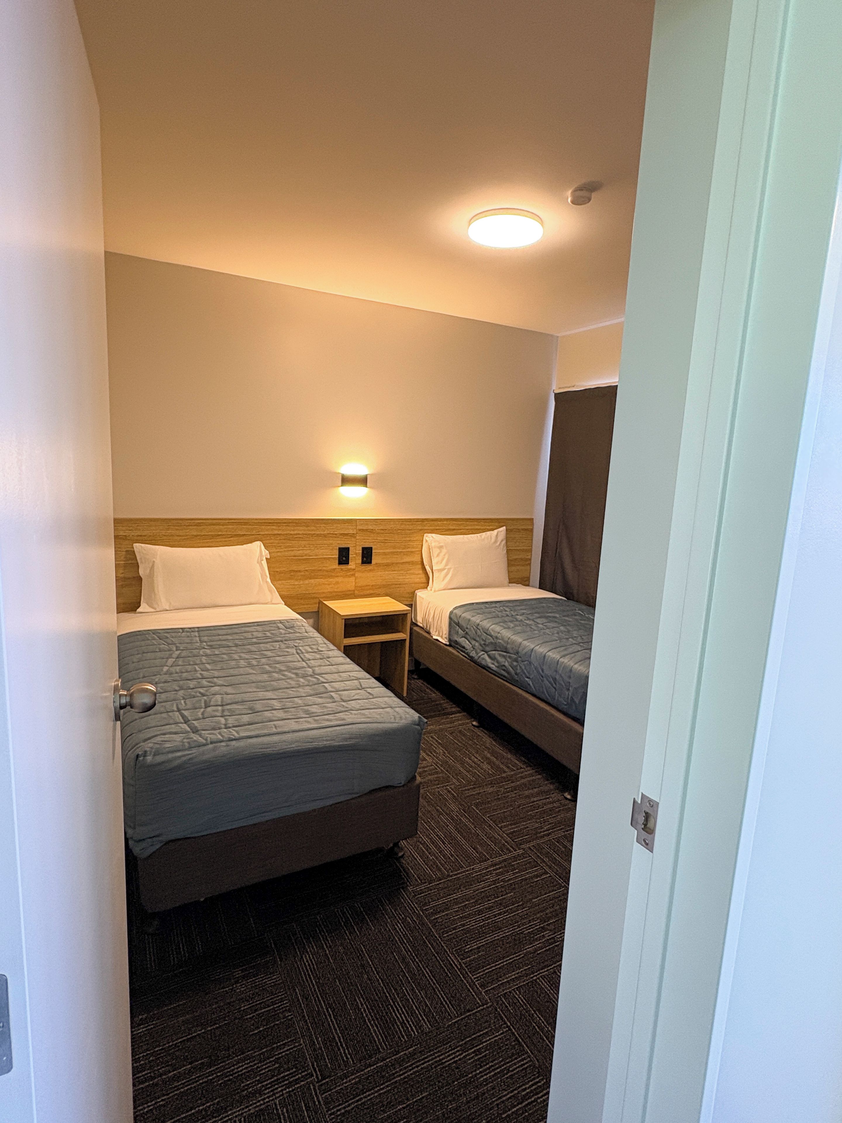Hotellihuone, jossa on kaksi sänkyä, yöpöytä, seinään kiinnitetty valaisin ja matolla peitetty lattia.