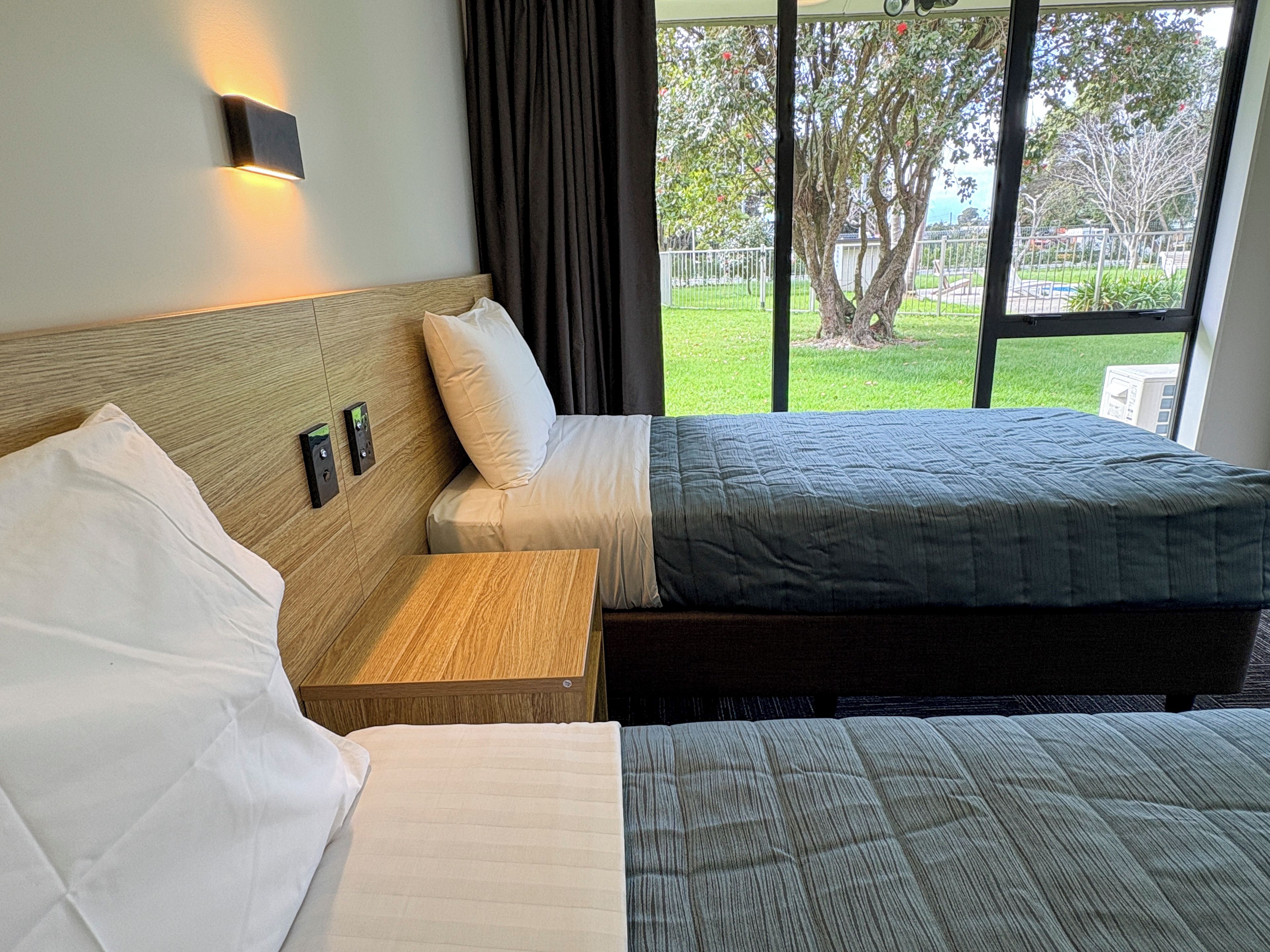 A hotel room with two beds, a nightstand, a wall-mounted light, and a view of the outdoors.