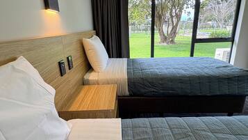 A hotel room with two beds, a nightstand, a wall-mounted light, and a view of the outdoors.