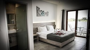 A modern hotel room with a large bed, a bathroom with a glass shower enclosure, a dining area with a view, and a decorative wall piece. - Vallian Village (Rhodes)