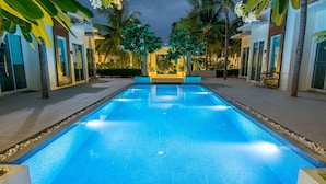 Una piscina con borde de azulejos, rodeada de palmeras y un edificio con ventanas amplias y una zona de estar.
