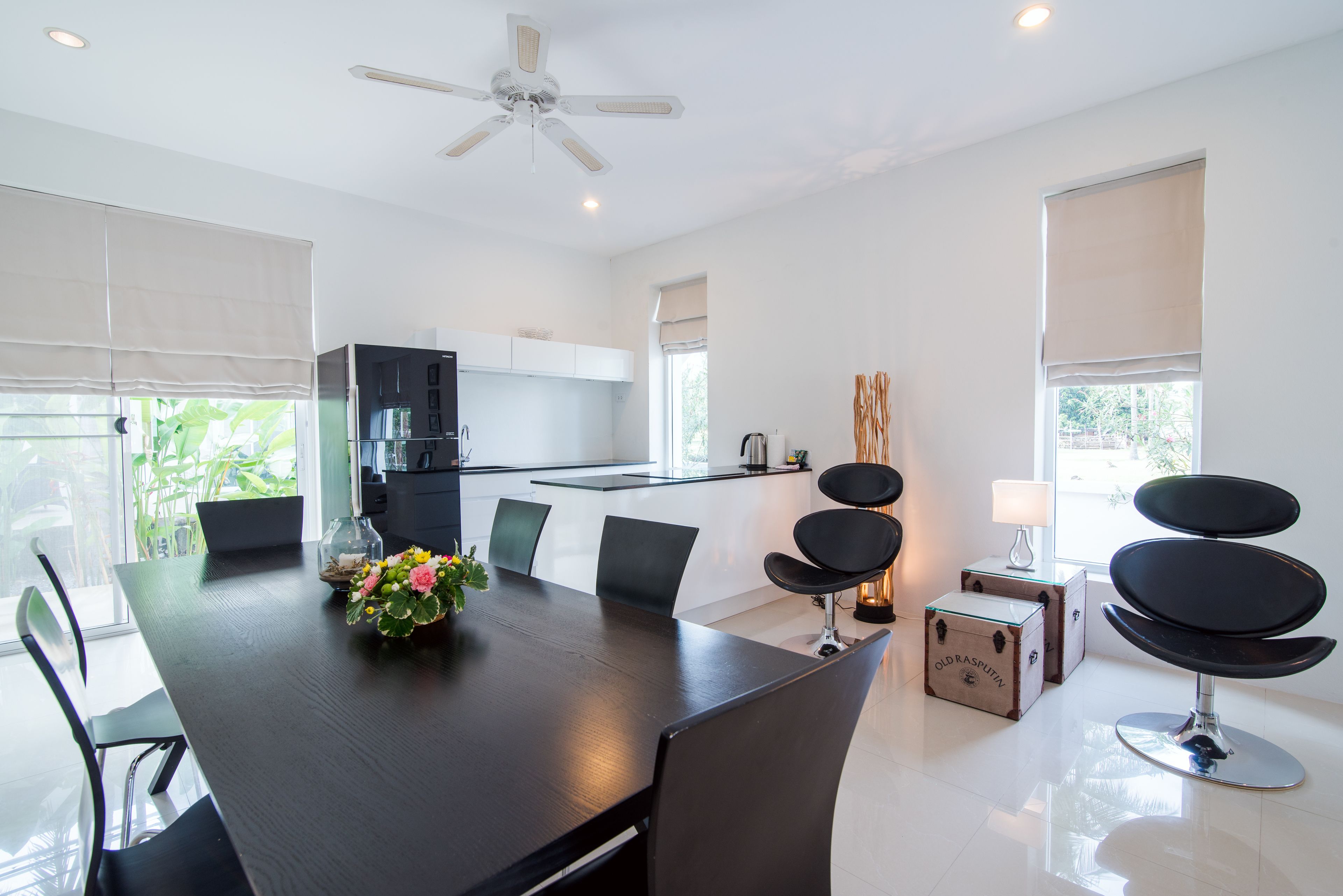 A modern dining area with a large table, chairs, a ceiling fan, and a kitchen in the background.