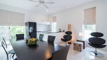 A modern dining area with a large table, chairs, a ceiling fan, and a kitchen in the background.