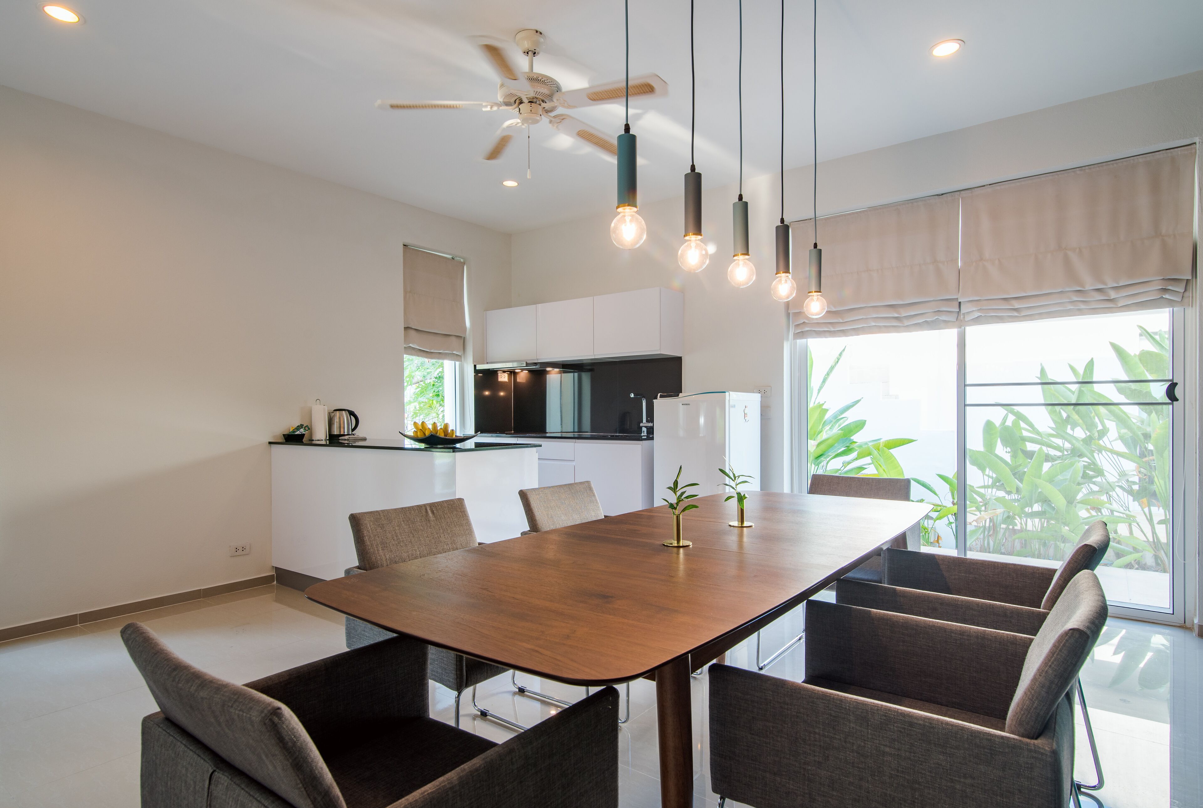 Un comedor moderno con una mesa y sillas de madera, un ventilador de techo, luces colgantes y vistas a la cocina y follaje exterior.