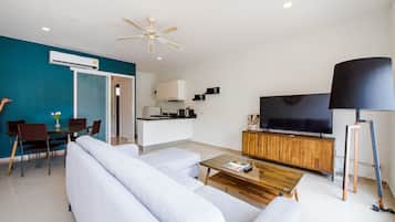 Ein modernes Wohnzimmer mit einer Couch, einem Couchtisch und einem Fernseher. Es gibt eine Küchenzeile mit Schränken und einem Deckenventilator.