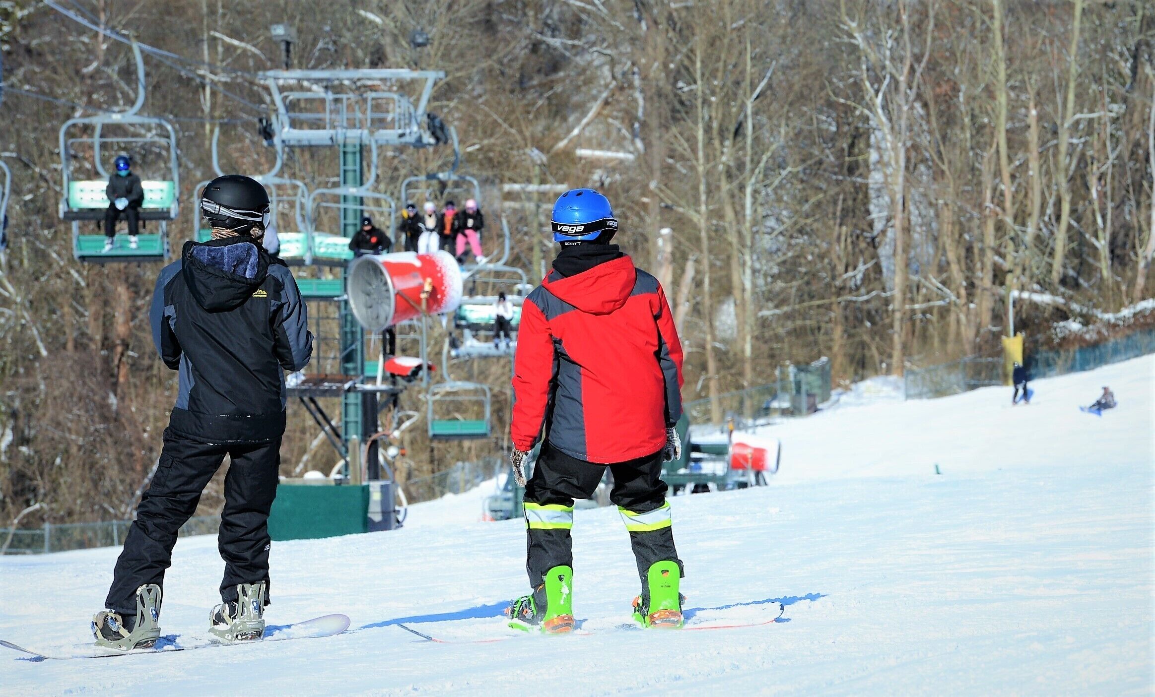 Twee personen staan op een besneeuwde helling, met skiliften op de achtergrond.