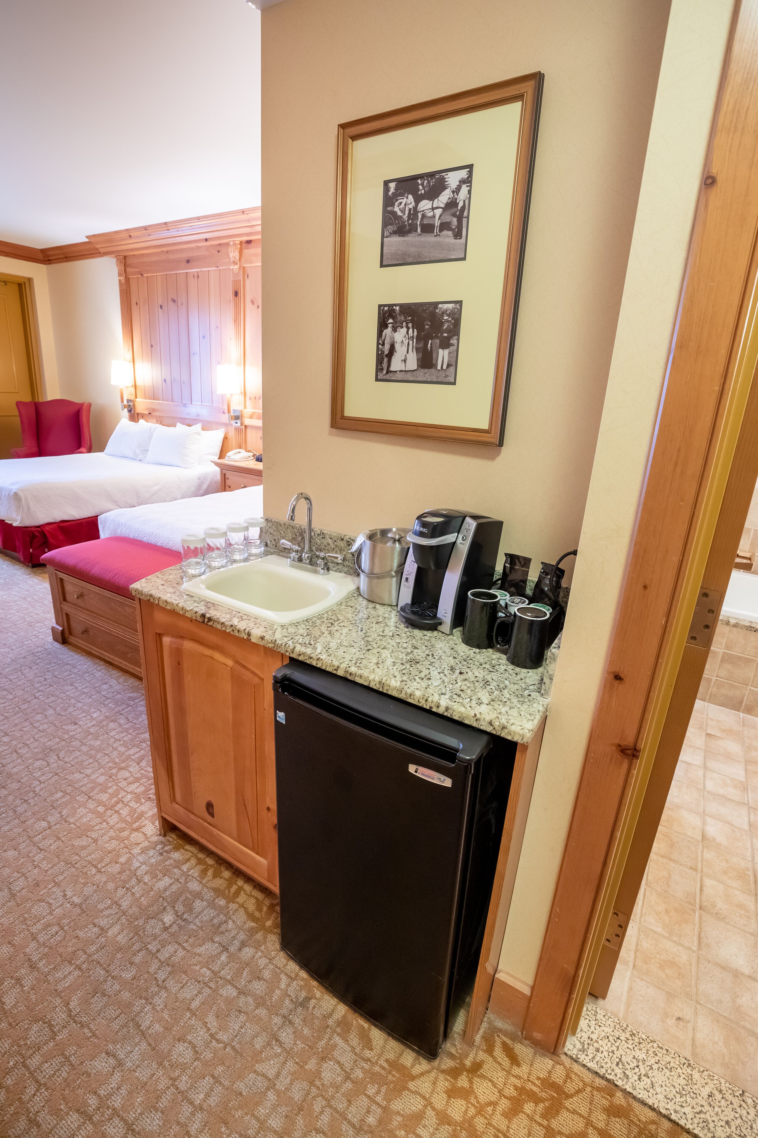 A compact hotel room with a kitchenette, a bed with red bedding, and a framed photo on the wall.
