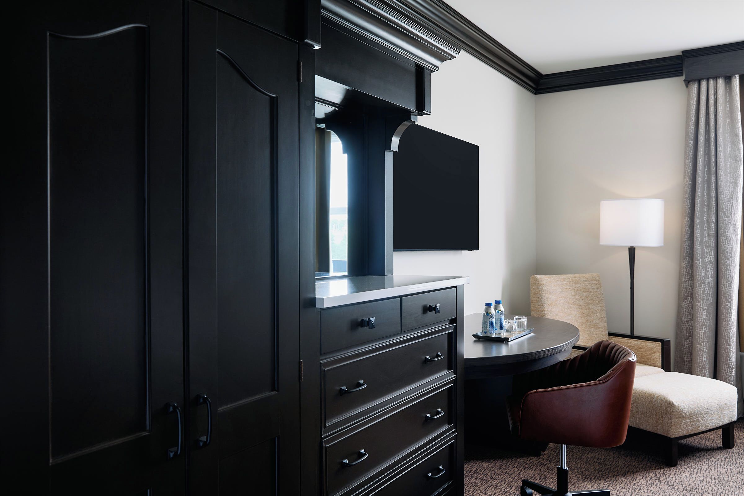 A hotel room with a dark wooden wardrobe, a flat-screen TV, a round table with a lamp, a chair, and a view of the outdoors.