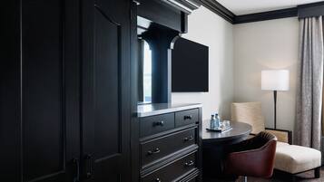A hotel room with a dark wooden wardrobe, a flat-screen TV, a round table with a lamp, a chair, and a view of the outdoors.