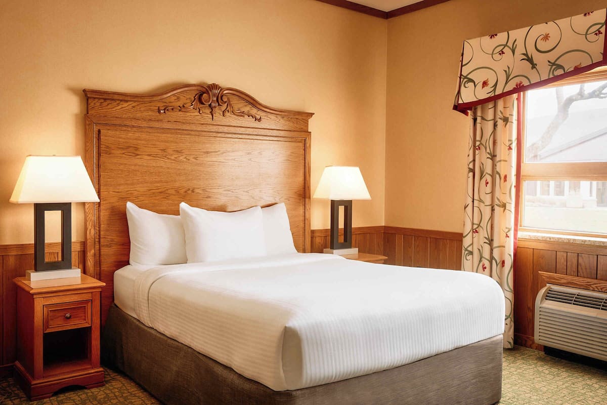 A neatly made bed with a wooden headboard, two bedside lamps, and a window with floral curtains.