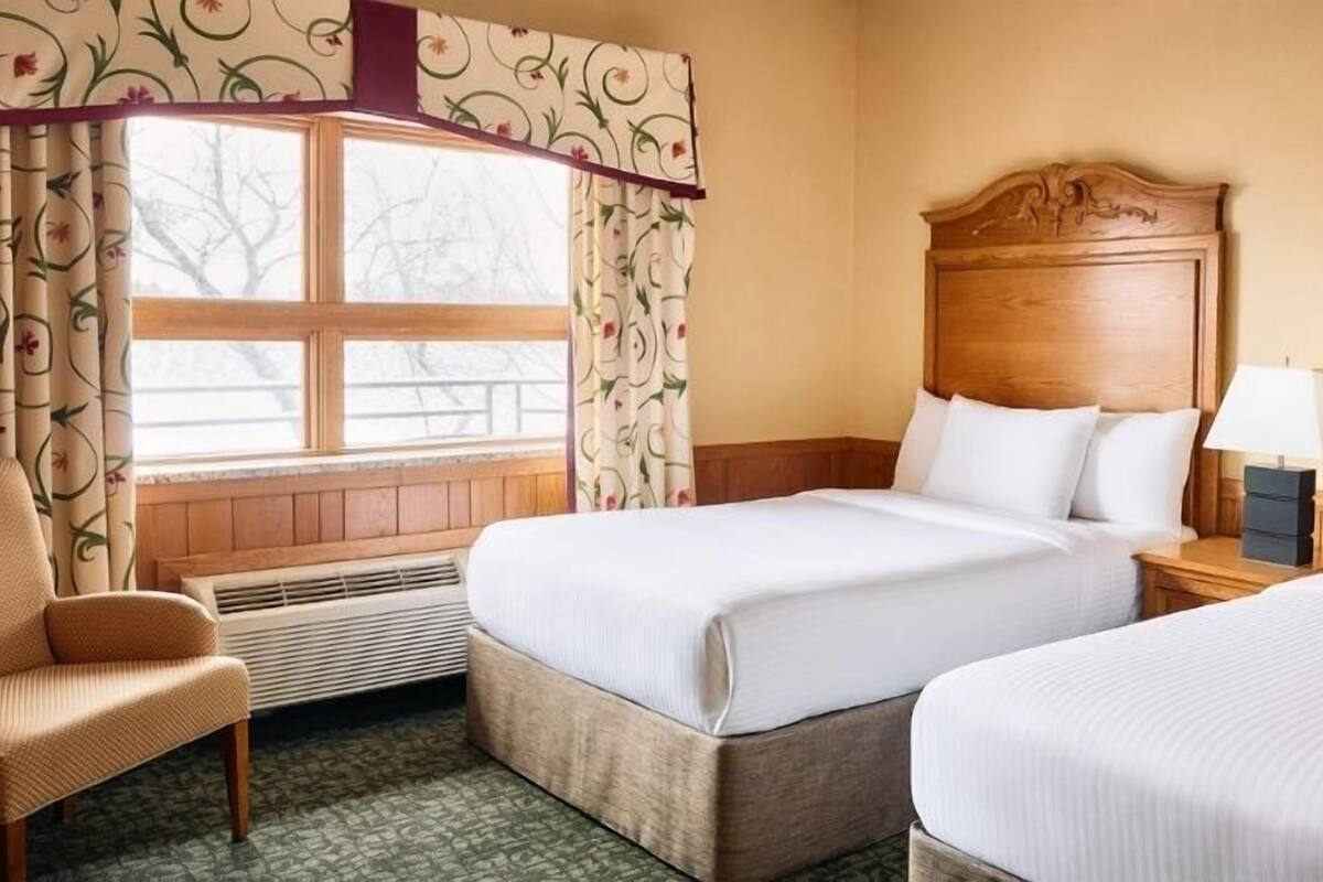 A hotel room with two beds, a chair, a nightstand with a lamp, and a window with curtains.