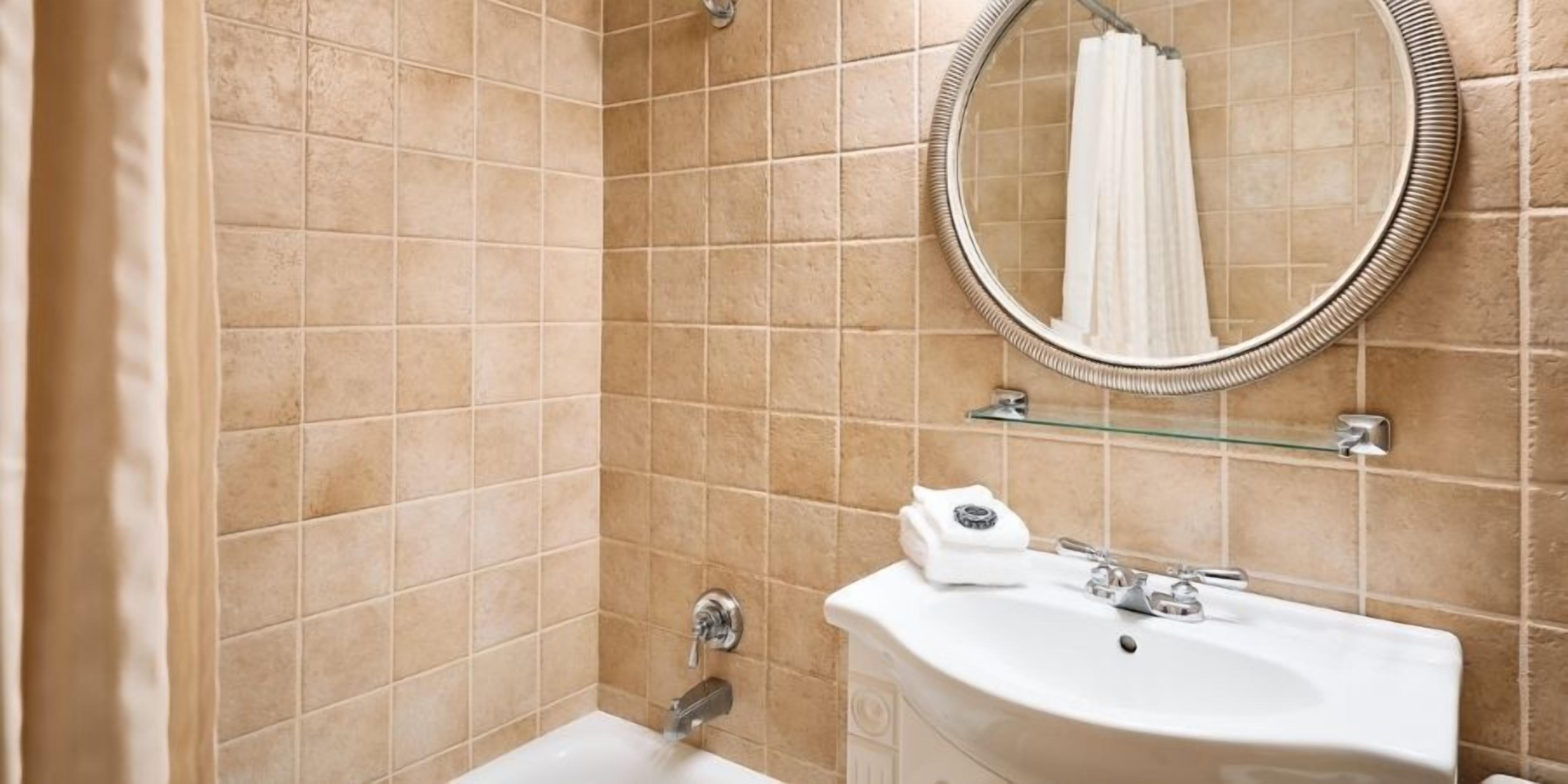 A bathroom with beige tiles, a white sink, a mirror, and a glass shelf.