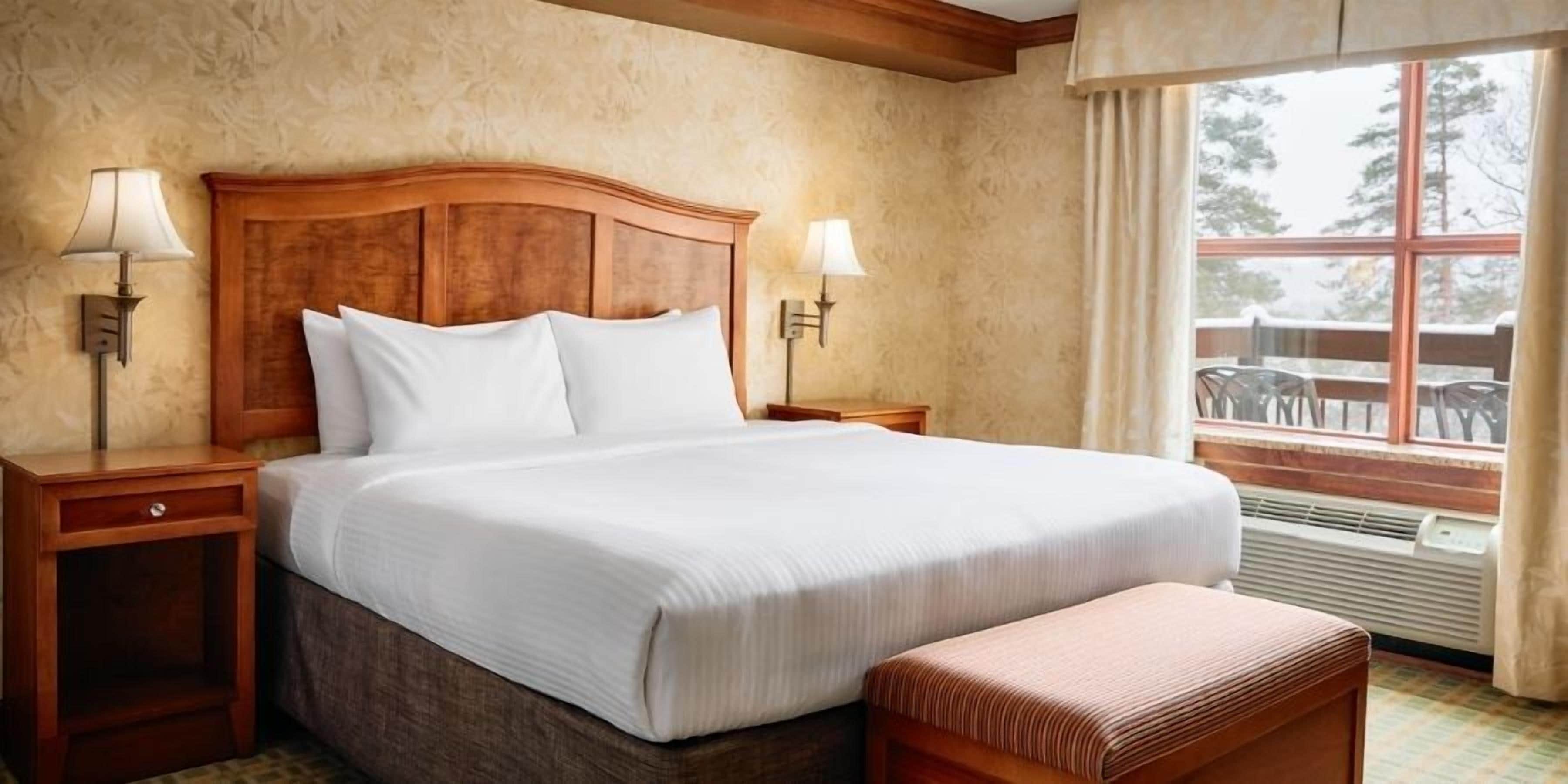 Hotelkamer met een groot bed, twee nachtkastjes, een bankje, een raam met uitzicht op bomen en een wand met een structuur.