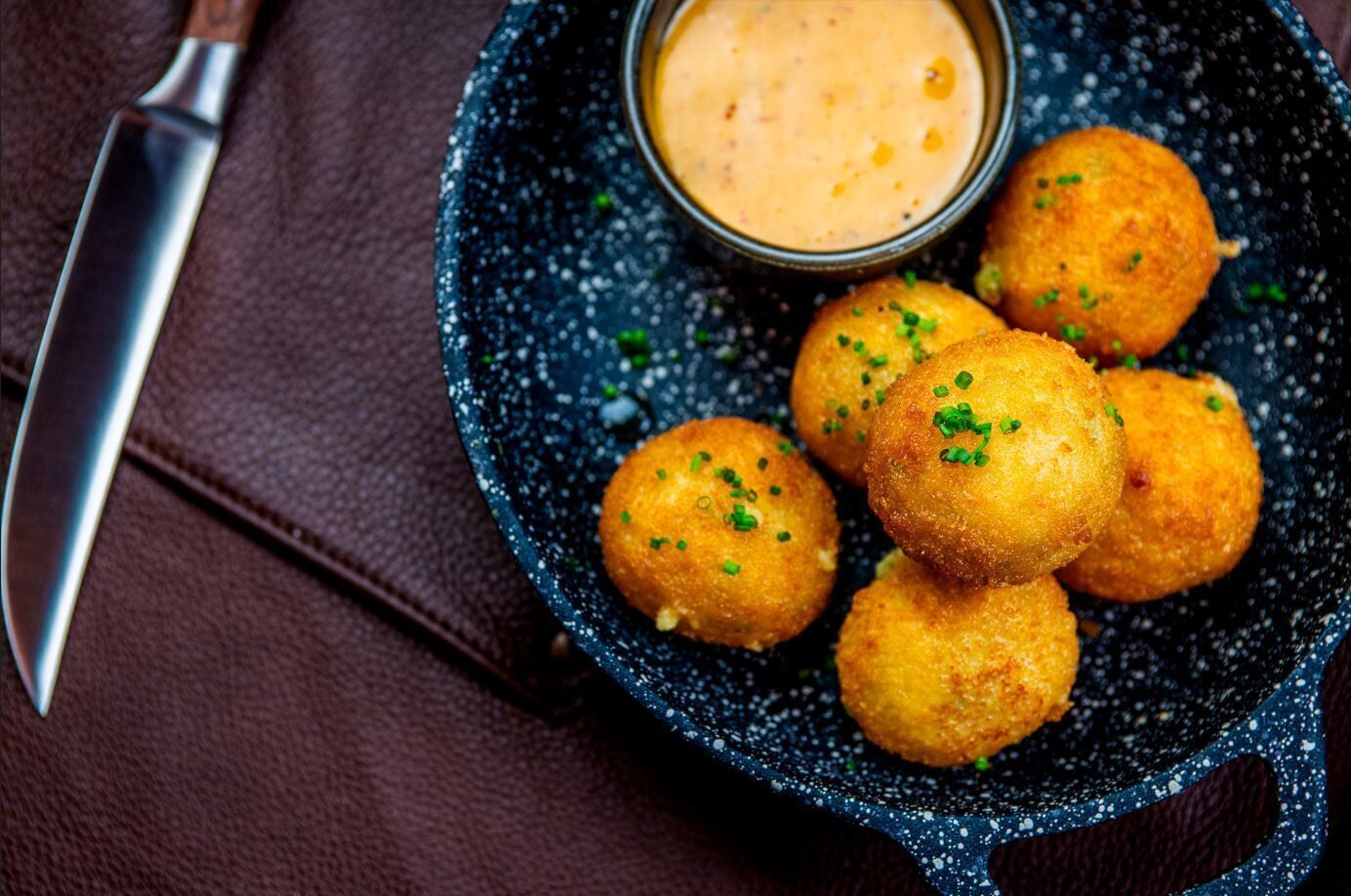 A plate of golden-brown croquettes with a dipping sauce, placed on a dark speckled plate.
