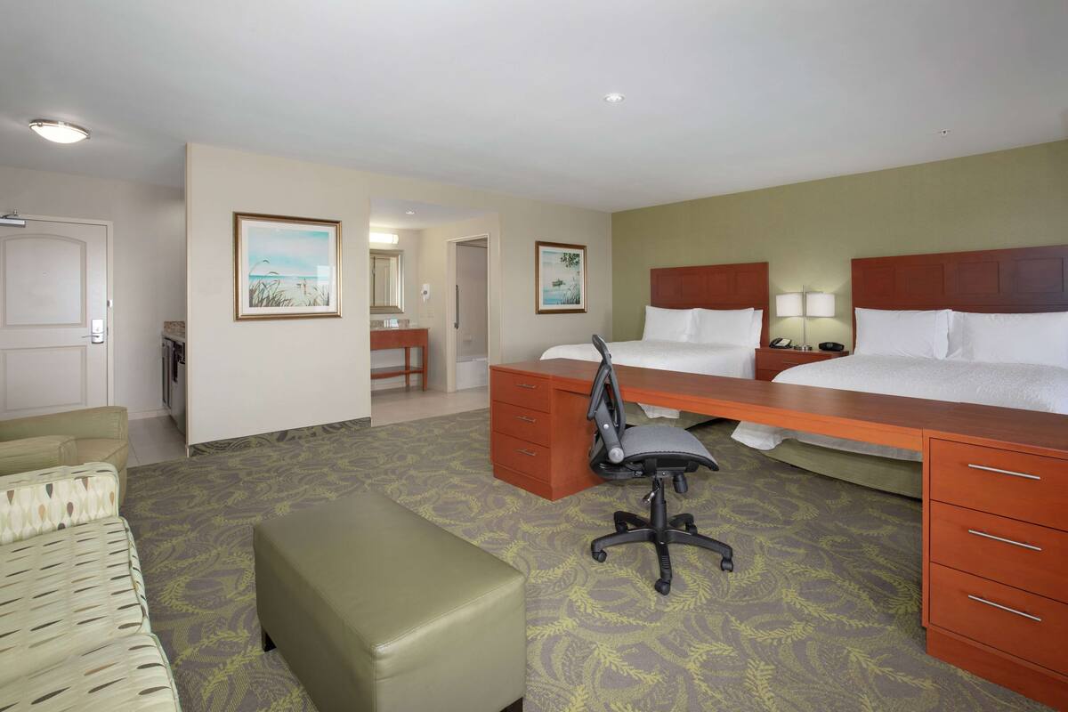 Une chambre d’hôtel avec deux lits, un bureau, une chaise, un petit canapé et une salle de bain visible à travers une porte ouverte.