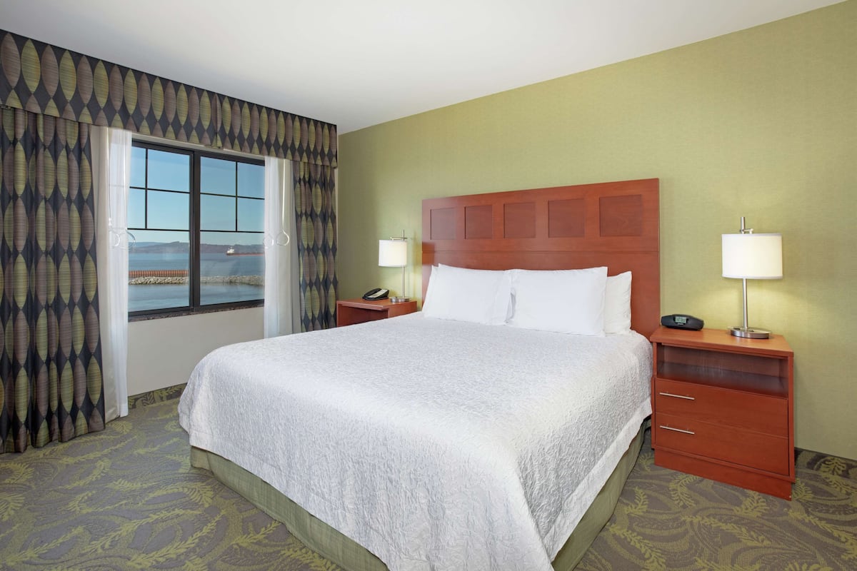 Une chambre d’hôtel avec un grand lit, deux tables de chevet, une petite table, une vue sur la mer et un rideau à motifs.