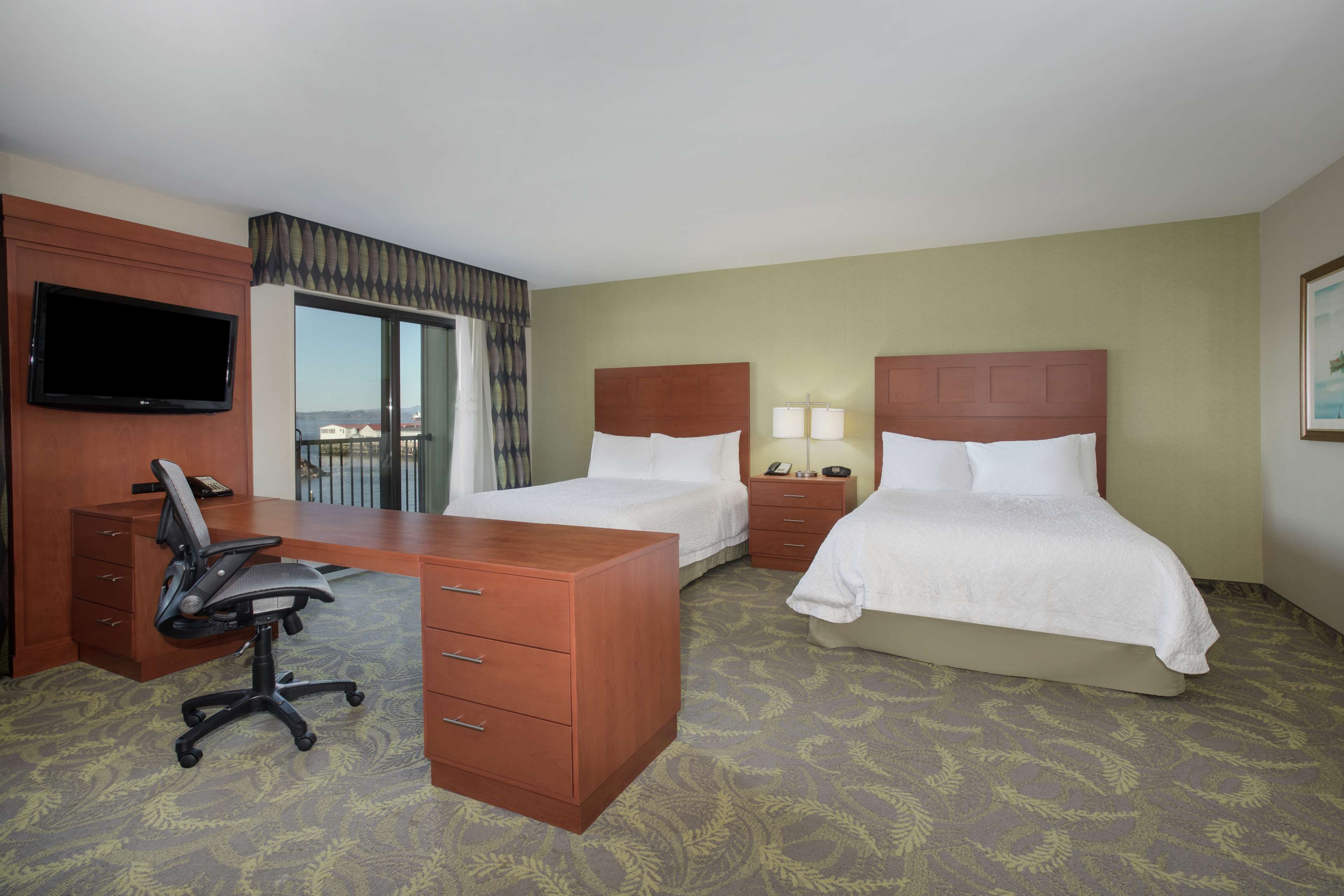 A hotel room with a desk, two beds, a TV, and a view of the outside.