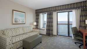 A hotel room with a sofa, a small table, a chair, a window with curtains, a radiator, and a framed picture on the wall. - Hampton Inn and Suites Astoria (Astoria)
