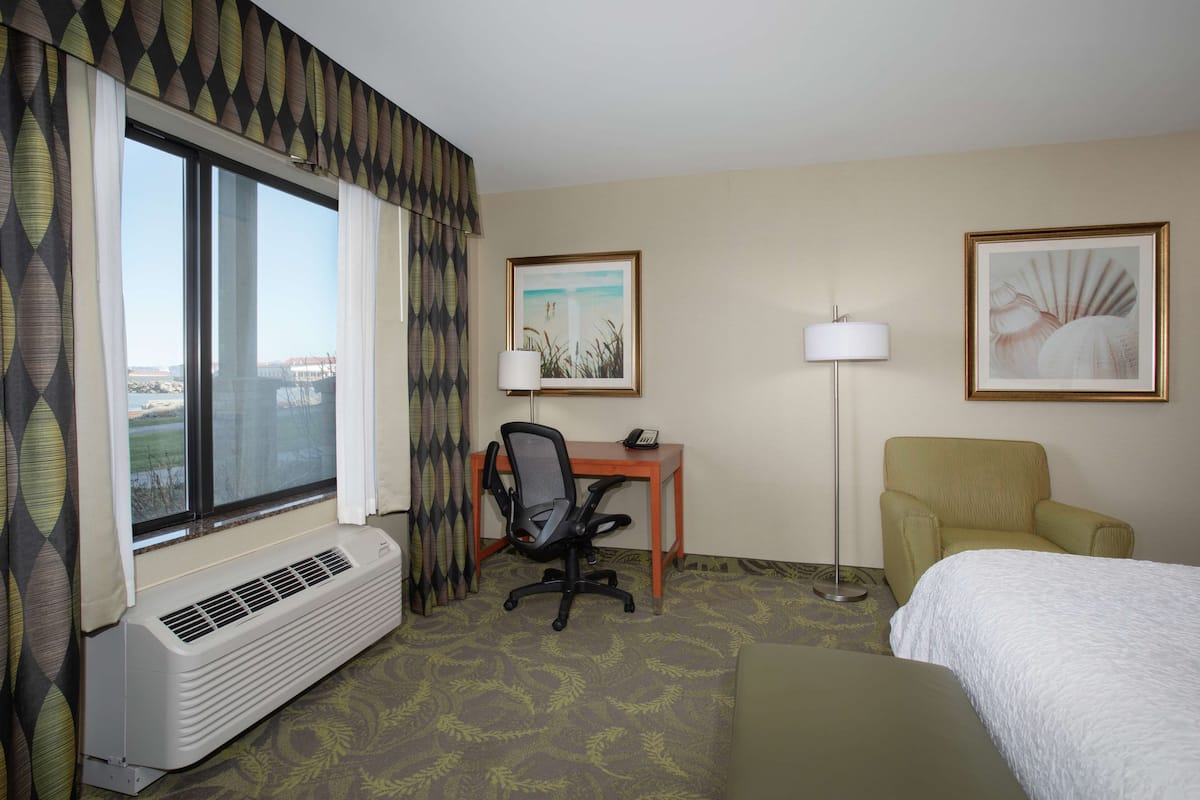 Une chambre d’hôtel avec un lit, un bureau et une chaise, une lampe sur pied et une grande fenêtre avec des rideaux.