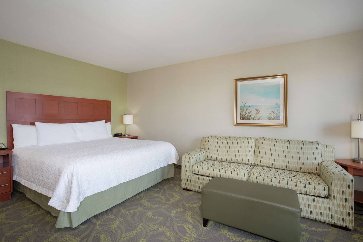 Une chambre d’hôtel avec un lit, un canapé, un pouf, une table de chevet et un tableau au mur.