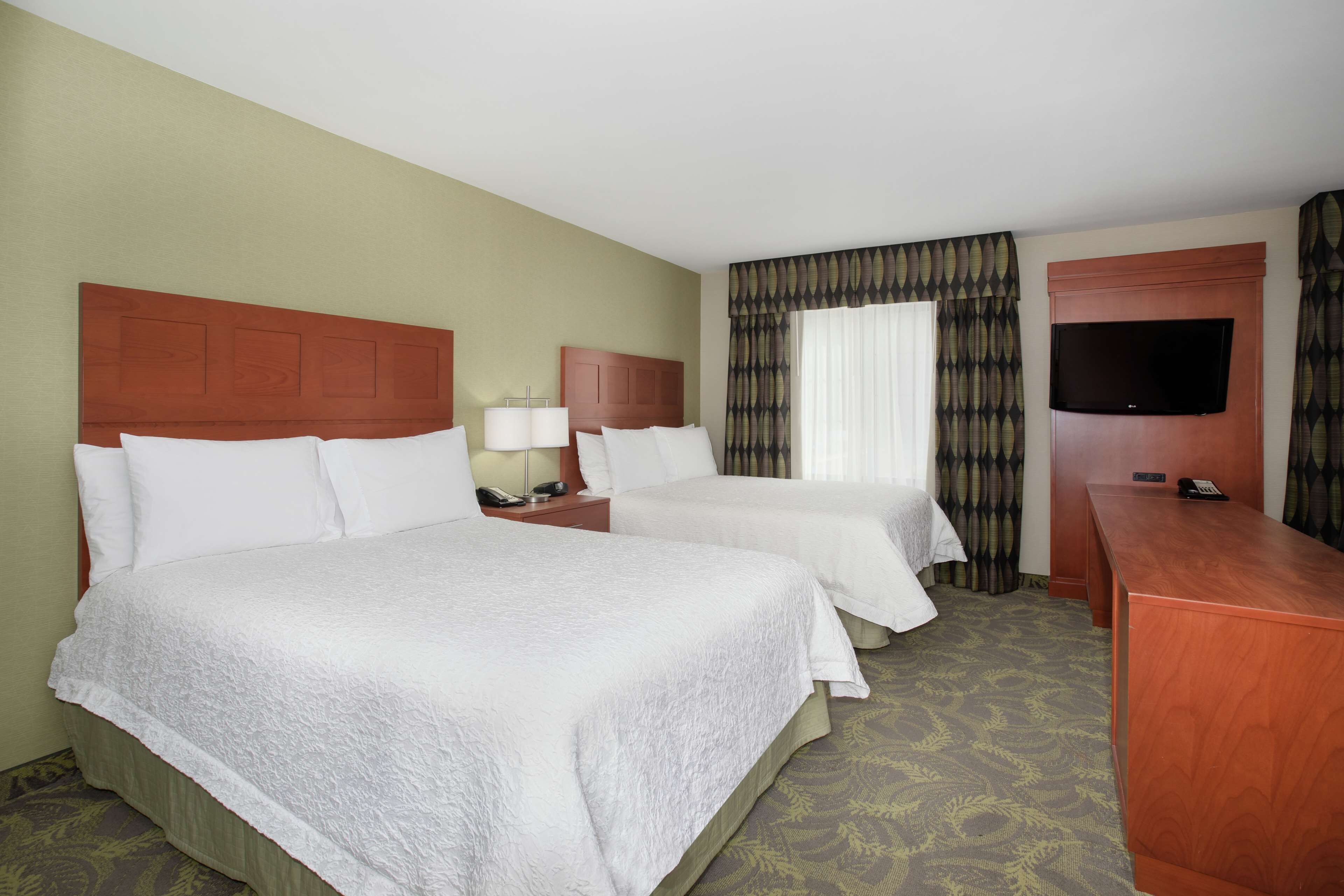 A hotel room with two beds, a TV, a desk, and a window with curtains.