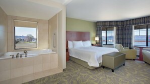 A hotel room with a large bathtub, a bed with a headboard, a chair, and a view of the city through the window. - Hampton Inn and Suites Astoria (Astoria)