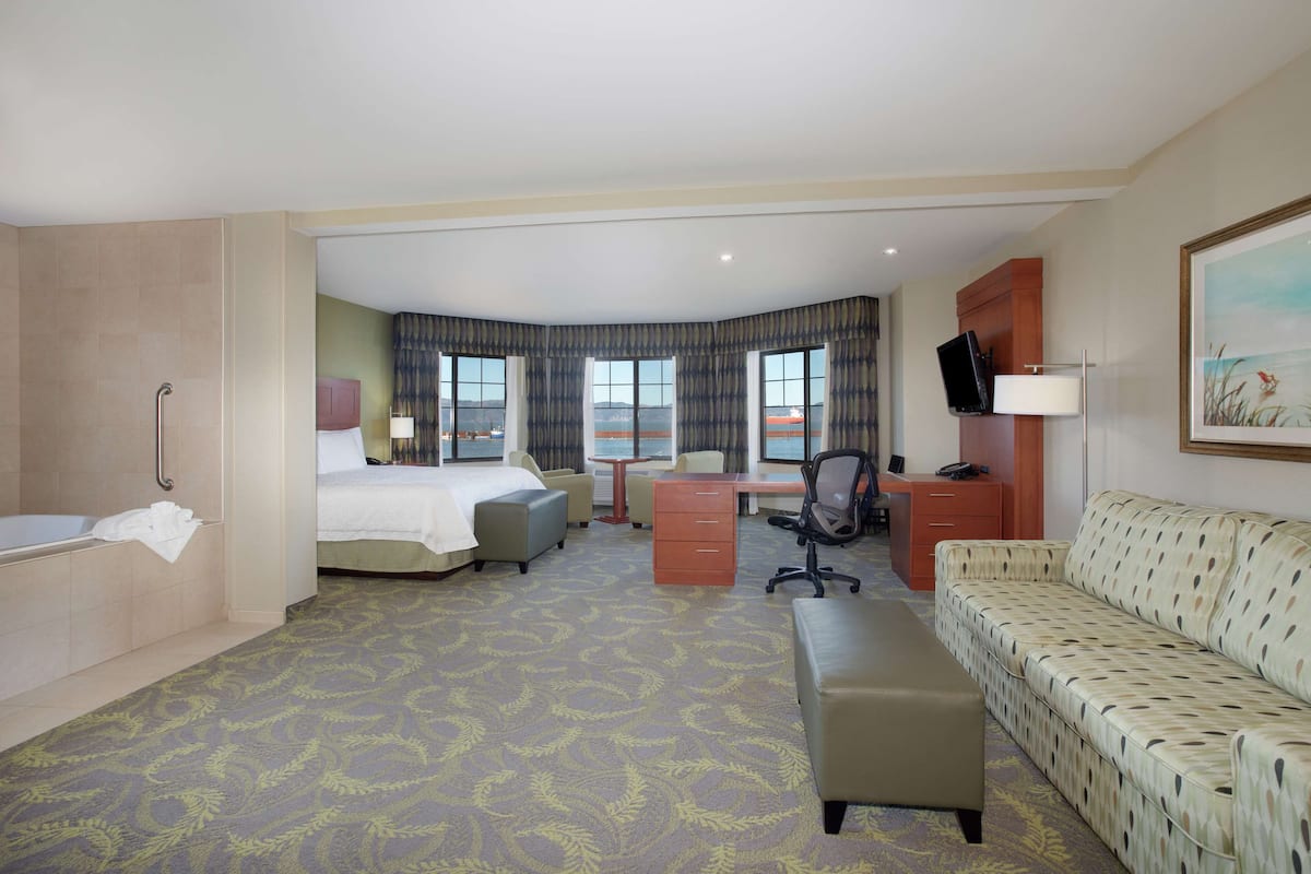 Une chambre d’hôtel comprenant un lit, un bureau, une chaise, un canapé, une salle de bain et une grande fenêtre donnant sur un paysage.