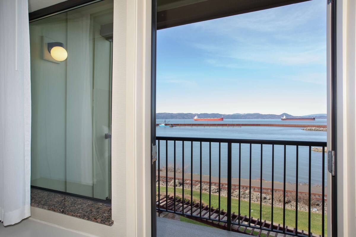 Un balcon offrant une vue sur une étendue d’eau paisible, une côte lointaine et un ciel dégagé.
