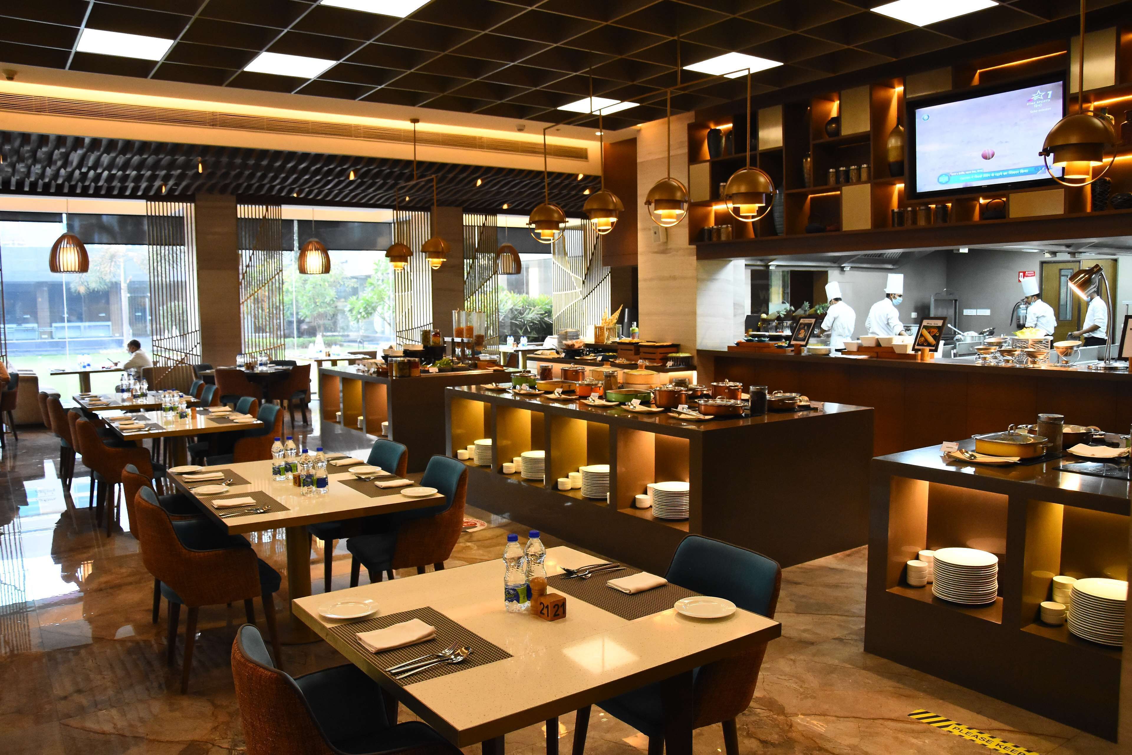 Un restaurant moderne doté de plusieurs tables, d’un espace cuisine et d’un grand écran diffusant du contenu.