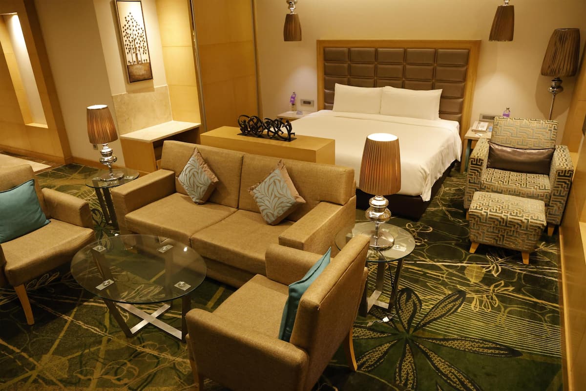Una habitación de hotel con una cama grande, una zona de estar con una mesa de vidrio y una alfombra estampada.